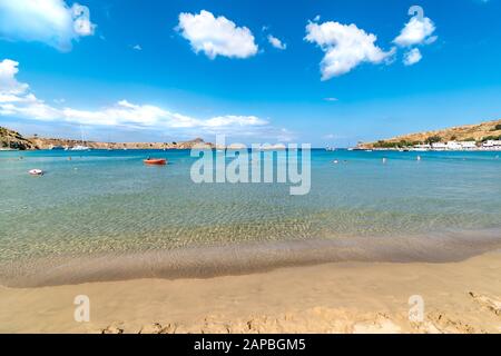 Blick auf den sandigen Strand und die Menschen im Meer in der Bucht von Lindos, Wolken am blauen Himmel (Rhodos, Griechenland) Stockfoto