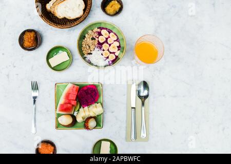 Kontinentales gesundes Frühstück mit Orangensaft, tropischem Obstteller, organischen Smoothies, Brotkorb, Marmelade und Butter auf Marmortisch mit Blumen. Draufsicht. Stockfoto