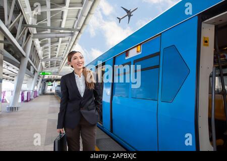 Junge, schöne asiatische Geschäftsfrau, die einen Sack hält und auf die Ankunft des Zuges am Skytrain der Flughafen-Bahnverbindung wartet. Stockfoto