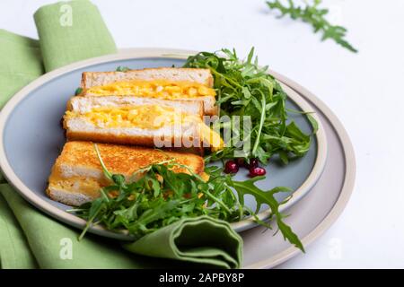 Sandwiches mit Rührei mit Käse und frischem Aurugulasalat, leckeres vegetarisches Frühstück auf weißem Tisch Stockfoto