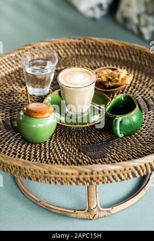Rattan Fach mit Cappuccino, Milch gravy Boat, Sugar Bowl, ein Glas Wasser und Cookies auf dem Blauen Sofa mit Kissen. Faul morgen Konzept. Stockfoto