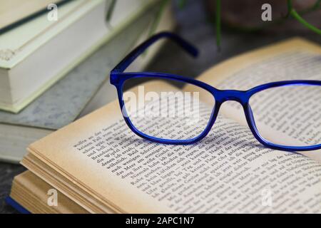 VIERSEN, DEUTSCHLAND - 21. AUGUST. 2019: Blick auf ein offenes gelobtes Buch mit blauer Lesebrille und Stapel alter Bücher auf runder Holztafel. Saftige Pflanze B. Stockfoto