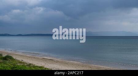 Flauschige graublaue Wolken treffen auf ruhiges dunkelblaues Meer weit von der sandigen Küste entfernt. Grünes Gras mit Blumen am Meer. Berge Hintergrund, Raum. Stockfoto