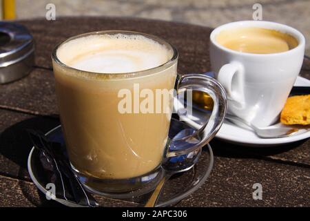 Nahaufnahme von isoliertem braunem holländischem Milchkaffee (Koffie verkeerd) in transparentem Glasbecher und weißem Espresso-Becher auf runder Holztafel Stockfoto