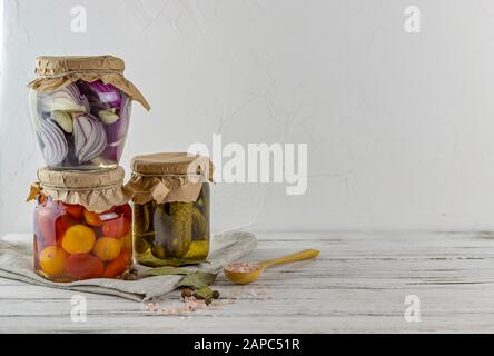 Drei Glasgefäße aus fermentierten Gurken, Tomaten und Zwiebeln. Gemüse auf hellem Hintergrund. Textil hellgrau. Gärung ist eine Quelle für Probi Stockfoto