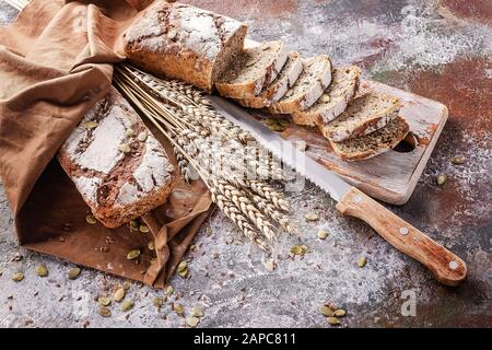 Frisch gebackenes Sauerteigsbrot mit Sonnenblumen und Kürbissamen an einer braunen Serviette. Brotscheiben auf Holzbrett und Küchenmesser. Weizenähren. Stockfoto