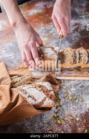 Weibliche Hände mit vertikalem Schuss schneiden frisch gebackenes Sauerteigsbrot mit Sonnenblumen- und Kürbissamen an einer braunen Serviette. Weizenähren. Stockfoto