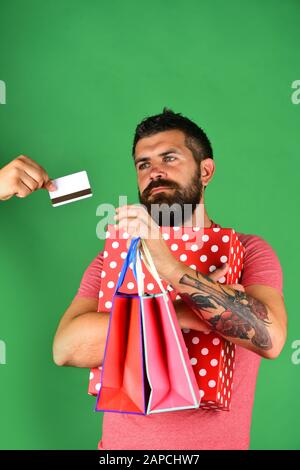 Mann mit Bart hält Einkaufstaschen und Schachtel. Geld- und Einkaufskonzept. Käufer hält große gestrichelte Schachtel mit Polka auf grünem Hintergrund. Kerl mit Bart und interessiertes Gesicht kauft ein. Stockfoto