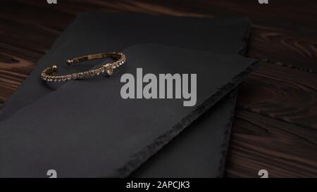 Golden mit Diamantarmband auf schwarzen Steinplatten. Goldenes Armband mit Rautenmuster auf groben, strukturierten Steinplatten auf Holztisch. Stockfoto