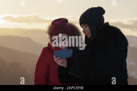 Zwei Mädchen im Winter tragen das betrachten von Bildern auf dem blauen Smartphone. Ein Mädchen, das ihre Freundschaftsfotos zeigt, die sie in den Bergen machen. Stockfoto