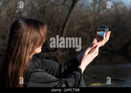 Frau mit dunkellangem Haar in schwarzen Roben hält polierte Klare Quarzkugel in ihren Händen vor dem See. Stockfoto