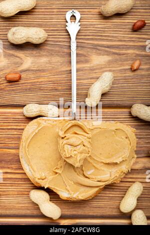 Vintage-Löffel mit cremiger Erdnussbutter auf einem Erdnussbutterbrot. Erdnüsse in der Schale und geschälte Erdnüsse auf braunem Holztisch. Flache Erdnusslage Stockfoto