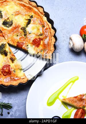 Vegetarische Pizza mit Tomaten, Brokkoli und Käse auf grauem Hintergrund. Traditionelles rustikales Küchenkonzept. Veganes Essen im Café oder Restaurant. Pizza Pastete in Backform aus Metall in der Nähe des Stücks auf Platte serviert Stockfoto