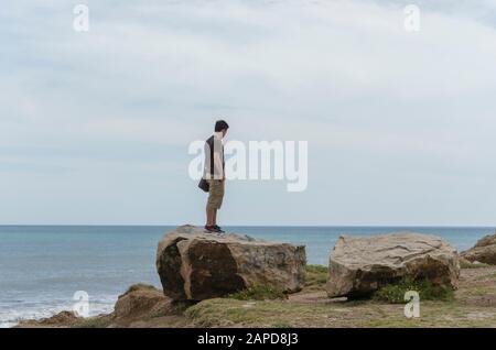 Junger Mann, der auf einem Felsen steht, der auf das Meer blickt, einen Sommernachmittag, in Miramar, Argentinien Stockfoto