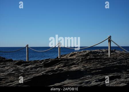 Das weiße Seil und die Pfosten umzäunen den Rand eines felsigen Felsens von Maroubra mit Blick auf das Meer und einen perfekten Horizont vom Rand Australiens. Stockfoto