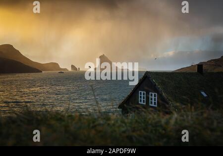 In Richtung der Rasenhäuser im Dorf Bøur zieht eine Schneedusche. Im Hintergrund werden Drangarnir und Tindhólmur von der Sonne beleuchtet. Faröer Stockfoto