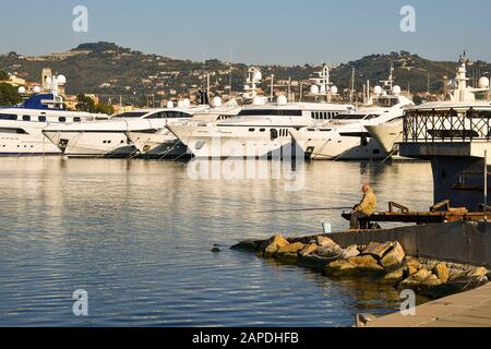 Der erfahrene Fischer fischt mit einer Stange im Hafen von Porto Maurizio mit Luxusyacht und der Küste der Blumenriviera im Hintergrund, Imperia Stockfoto