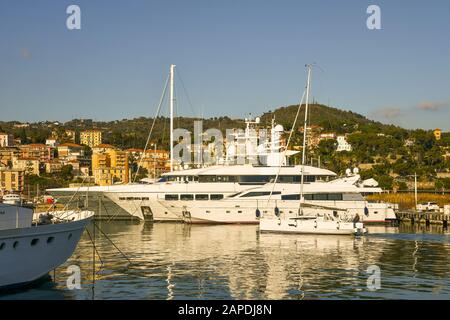 Ein Segelboot, das mit Luxusyachten am Dock und der Küstenstadt im Hintergrund den Hafen betritt, Porto Maurizio, Imperia, Ligurien, Italien Stockfoto