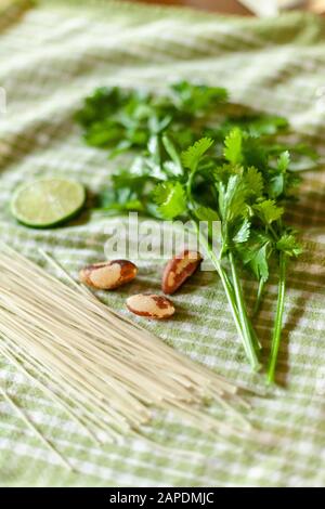 Die Zutaten für Cilantro Pesto sind auf einem grünen Gingham-Tuch angeordnet: Cilantro, brasilianische Nüsse und glutenfreie weiße Reisnudeln. Stockfoto