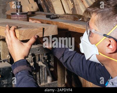 Ein Zimmermann, ein Handwerker in einer Atemschutzmaske und eine Baubrille, wählt Holzplatten für und verarbeitet die Rohlinge seiner Produkte Stockfoto