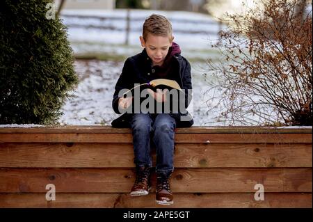 Kleiner Junge, der auf Holzbrettern sitzt und die bibel liest In einem im Schnee bedeckten Garten Stockfoto