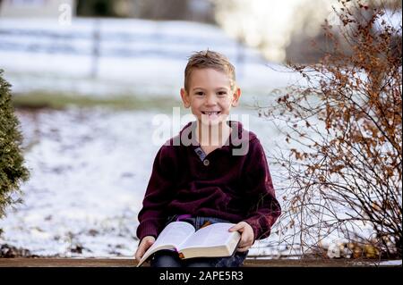 Kleiner Junge, der auf Holzbrettern sitzt und die bibel liest In einem im Schnee bedeckten Garten Stockfoto