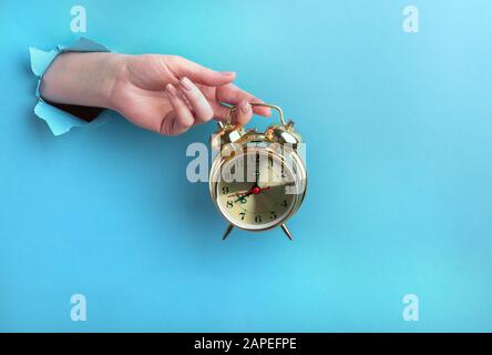 Weibliche Hand hält einen goldenen Wecker durch ein Loch auf blauem Grund. Zeitkonzept mit Platz für Text. Stockfoto