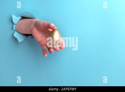Goldenes Ei in einer weiblichen Hand auf blauem Grund mit einem Loch. Horizontale Ausrichtung mit Kopierbereich. Stockfoto