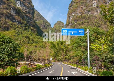 Leere Straße gegen Karsthügel und Vegetation an einem sonnigen Tag in Yangshuo mit blauem Straßenschild mit Yangshuo und Gaotian in chinesischen und rumänischen Schriftzeichen geschrieben... Stockfoto