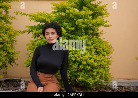 Frau mit lockigen Haaren, die im Hintergrund mit grünem Baum sitzt Stockfoto