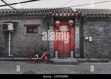 Tür rot lackiert und für das chinesische Neujahr dekoriert, in einem Hutong, Peking, China. Mobike Fahrrad vor dem Hotel geparkt. Startseite/Handel Stockfoto
