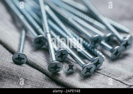 Eine Handvoll metallischer Selbstschneiderschrauben liegt vor dem Hintergrund eines alten Denimstoffs. Selektive Fokusaufnahme mit flachem Freiheitsgrad. Extreme Nahaufnahme. Stockfoto