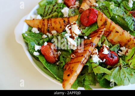 Schließen Sie einen Teller grünen Salats mit gerösteten Birnen und frischen Erdbeeren auf weißem Tisch Stockfoto