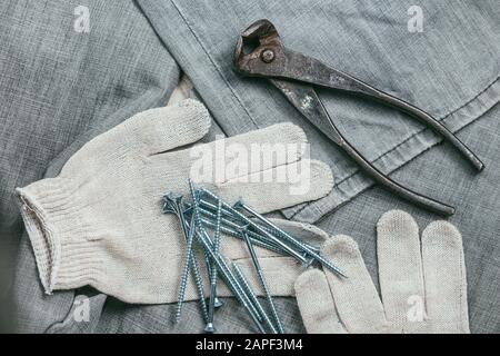 Eine Handvoll metallischer Selbstschneiderschrauben und sehr gealterte und rostige Zangen liegen auf Safety Strickhandschuhen für den industriellen Einsatz vor dem Hintergrund Stockfoto