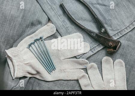 Eine Handvoll metallischer Selbstschneiderschrauben und sehr gealterte und rostige Zangen liegen auf Safety Strickhandschuhen für den industriellen Einsatz vor dem Hintergrund Stockfoto