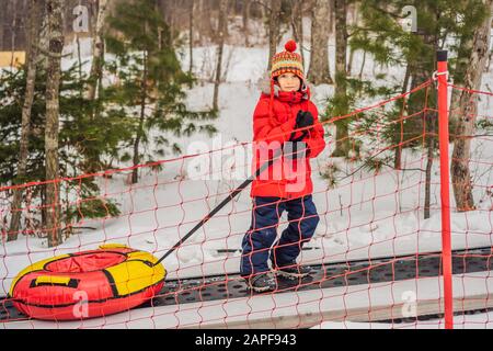 Junge mit Schlauch steigt auf einem Travelator auf den Berg. Kind hat Spaß an der Schneeröhre. Junge reitet einen Schlauch. Winterspaß für Kinder Stockfoto