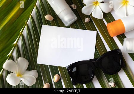 Sommerschablone mit Grußkarte, Sonnenbrille, tropischem Palmblatt, Sonnenschutzmittel, Sonnenschutzmittel und Blumen auf weißem Hintergrund Stockfoto