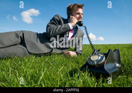 Fröhlicher Geschäftsmann, der sich auf einem sonnigen Flecken von hellgrünem Gras entspannt und mit einem altmodischen Drehwahltelefon spricht Stockfoto