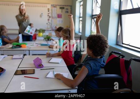 Gruppe von Schulkindern, die an Schreibtischen sitzen und die Hände in einem Schulunterricht heben Stockfoto