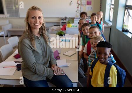 Schullehrerin, die in einem Klassenzimmer mit einer Gruppe von Schulkindern auf einem Schreibtisch sitzt Stockfoto