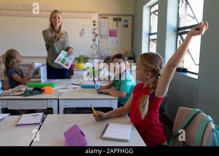 Schulmädchen sitzt an einem Schreibtisch und hebt ihre Hand in einem Schulklassenzimmer Stockfoto