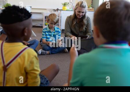 Schulleiterin sitzt quer auf dem Boden im Kreis mit einer Gruppe von Schulkindern Stockfoto
