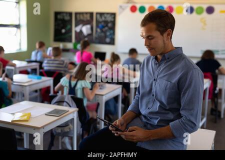 Männliche Schullehrerin, die auf einem Schreibtisch in einem Schulunterricht sitzt Stockfoto