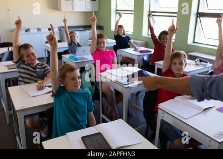 Gruppe von Schulkindern, die an Schreibtischen sitzen und ihre Hände heben, um zu antworten Stockfoto