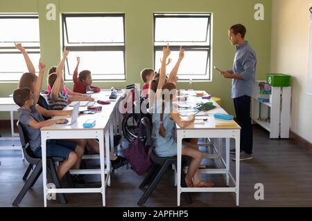 Gruppe von Schulkindern, die an Schreibtischen sitzen und ihre Hände heben, um zu antworten Stockfoto