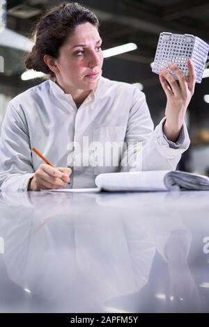 Brünette Frau in weißem Mantel mit Metallwürfel sitzt im Labor auf dem Tisch, verschwommener Hintergrund Stockfoto