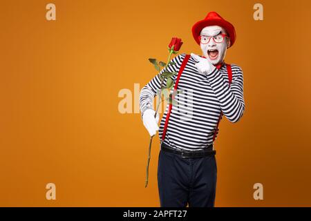 Fröhlicher MIME mit weißem Gesicht in rotem Hut und gestreiftem T-Shirt mit Rose auf leerem orangefarbenem Hintergrund im Studio Stockfoto