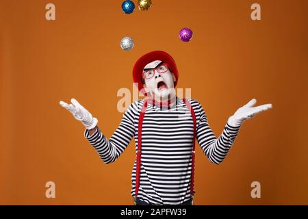 MIME man in Weste und Red hat jongliert mit bunten Bällen auf orange leerem Hintergrund Stockfoto