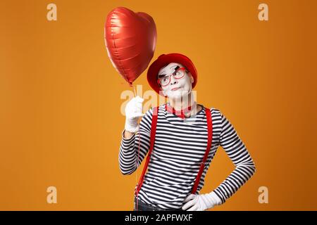 Ernster MIME Clown in rotem Hut und Weste hält Herzball in den Händen auf leerem orangefarbenem Hintergrund Stockfoto