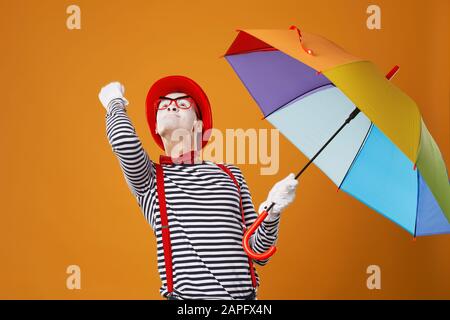 MIME, der mit einem mehrfarbigen Regenschirm in der Hand nach oben blickt und im Studio die Faust isoliert auf leerem orangefarbenem Hintergrund zeigt Stockfoto